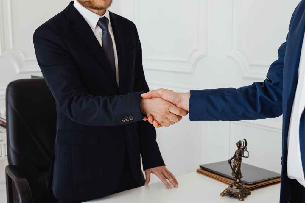 Two men shaking hands in a white room with a statue of the scales of justice on the desk beneath them